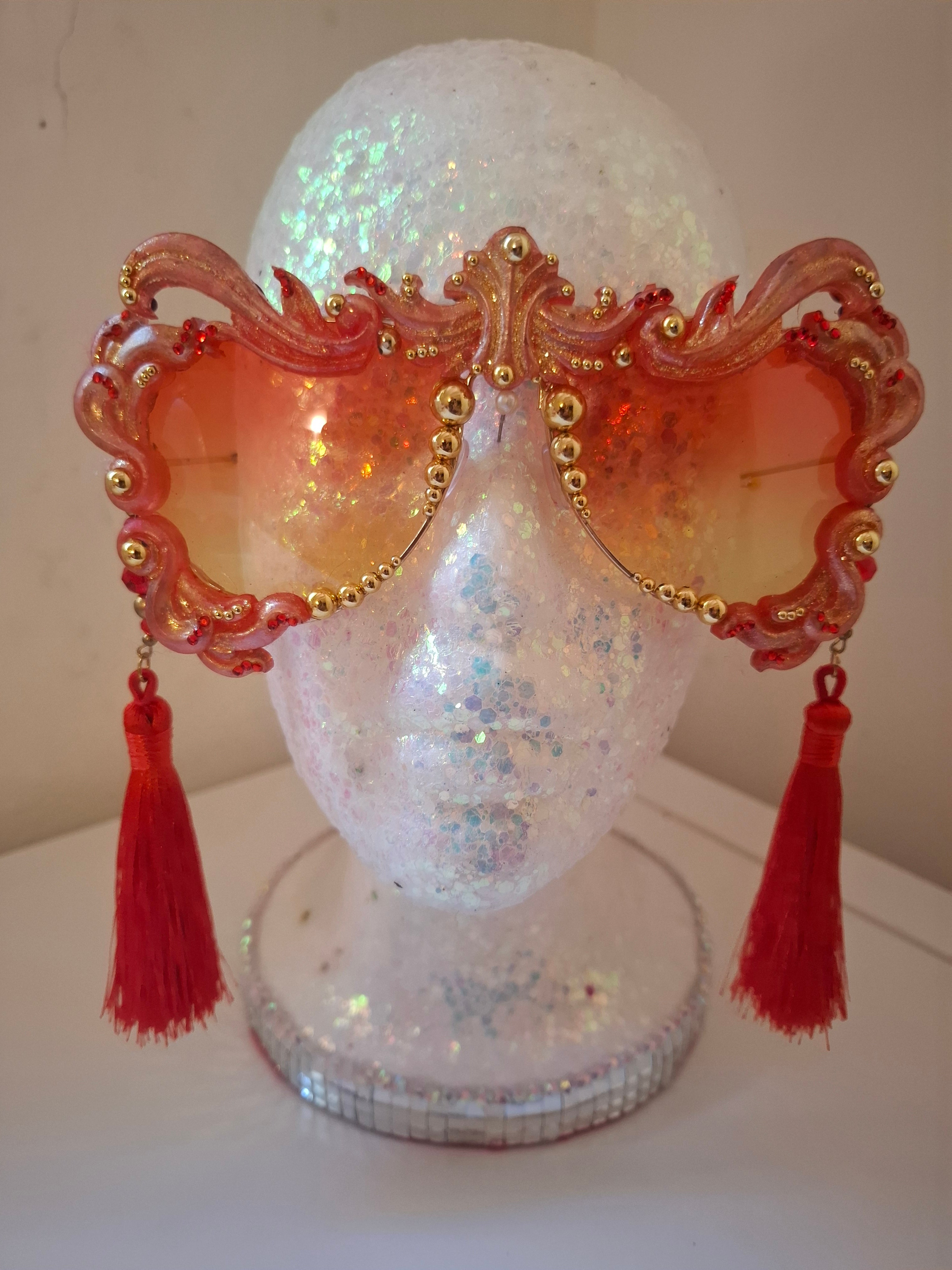 Hand-sculpted sunglasses in coral and gold flame scrollwork with crimson tassels. 