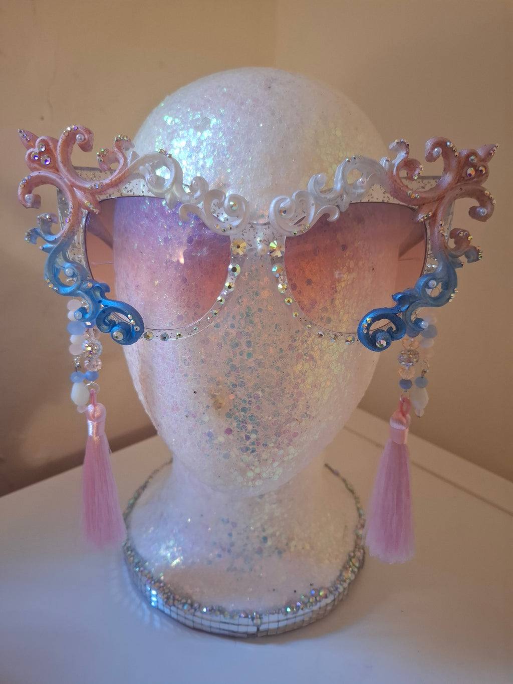 Hand-sculpted Pride sunglasses in trans flag colors — pink, white, and blue, with beaded tassels.
