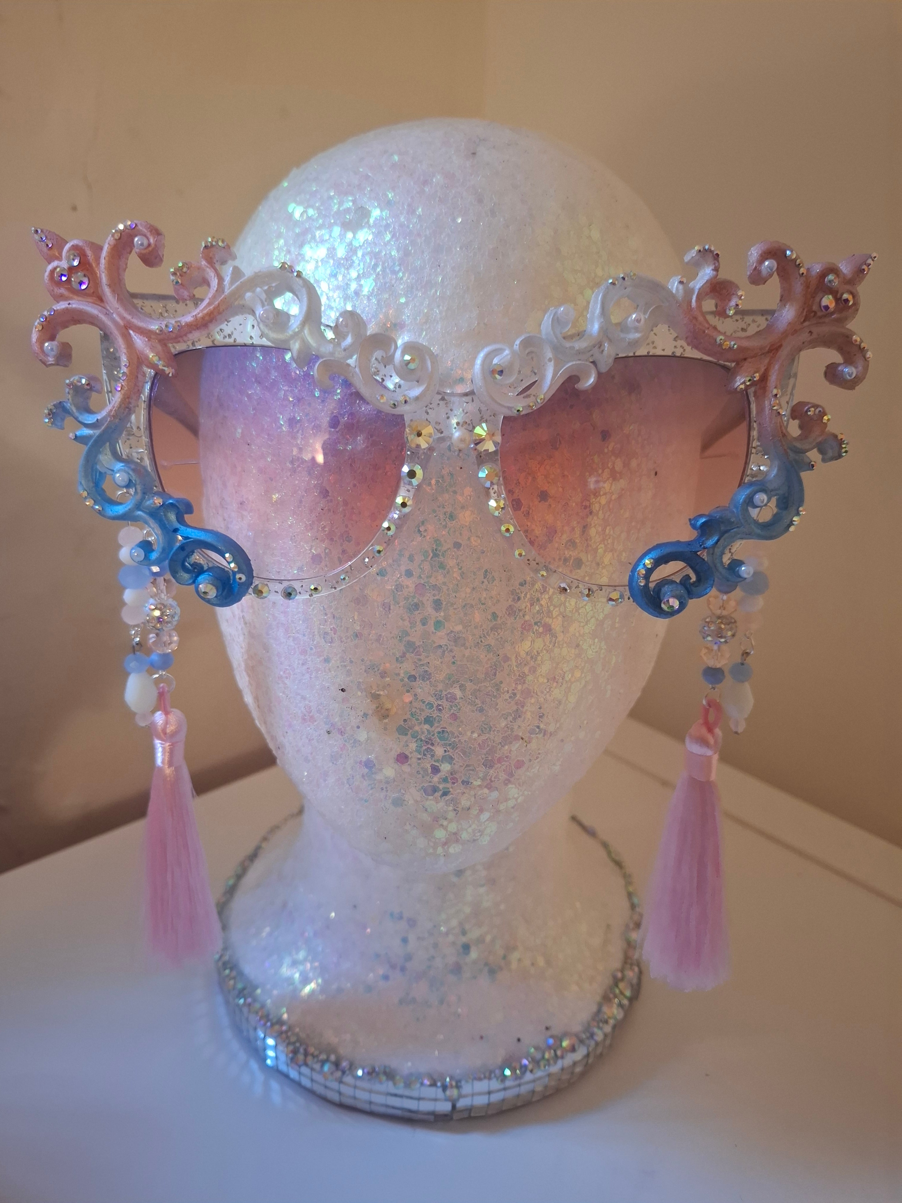Hand-sculpted Pride sunglasses in trans flag colors — pink, white, and blue, with beaded tassels.
